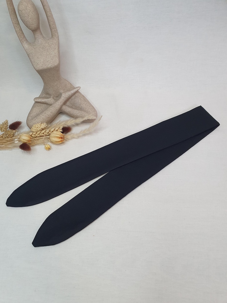Puede incluir: Un cintur&oacute;n de tela negra con extremos puntiagudos, sobre una superficie blanca. El cintur&oacute;n es largo y parece estar hecho de un material liso. Una estatua beige de una persona en postura de yoga est&aacute; en el fondo, junto con flores secas.