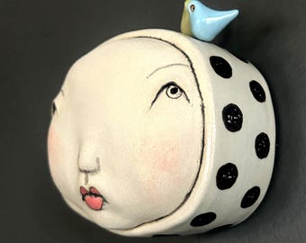 May include: A whimsical ceramic sculpture of a face with a polka-dotted head covering and a small blue bird perched on top. The face has a serene expression with pink lips and cheeks. The sculpture is off-white with black polka dots.