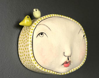 May include: A ceramic wall art piece featuring a stylized face with closed eyes and a small, pursed mouth. The face is cream-colored with pink cheeks and red lips. Two small yellow birds perch on top of the head, which is adorned with a yellow dotted pattern.