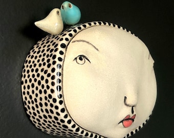 May include: A ceramic sculpture of a crescent moon face with a black and white polka dot pattern. Two small bird figurines, one white and one blue, sit atop the moon. The face has a pink lip color and is set against a black background.