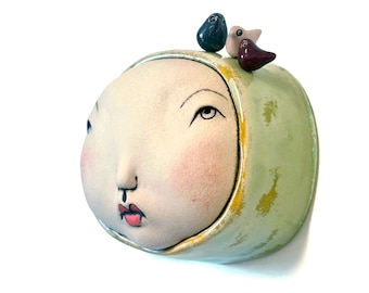 May include: A ceramic sculpture of a face with a light green hood and three small bird figurines on top. The face has rosy cheeks, red lips, and delicate black line details. The birds are dark blue, pink, and purple.