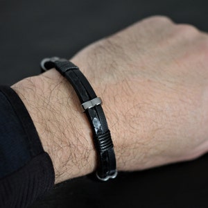 K&ouml;nnte beinhalten: Ein schwarzes Lederarmband mit einem silbernen Verschluss. Das Armband ist geflochten und hat eine strukturierte Optik.