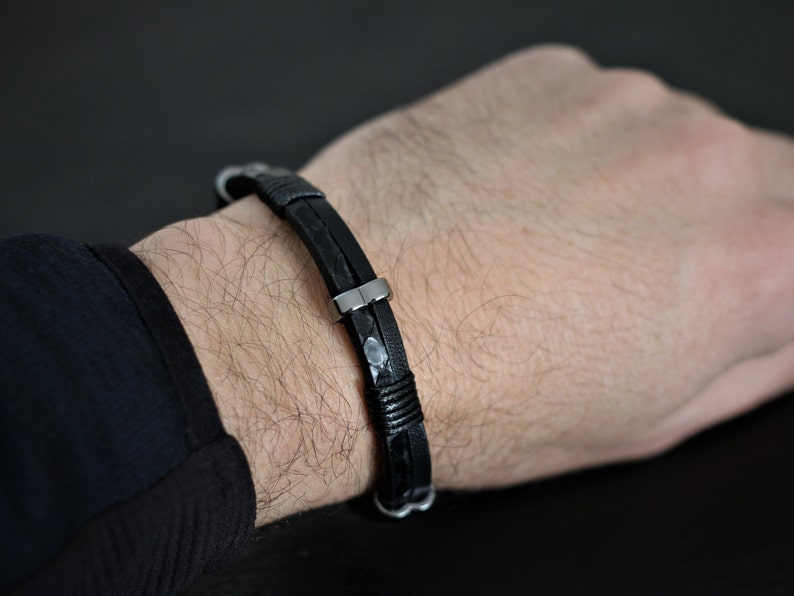 K&ouml;nnte beinhalten: Ein schwarzes Lederarmband mit einem silbernen Verschluss. Das Armband ist geflochten und hat eine strukturierte Optik.