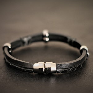 K&ouml;nnte beinhalten: Ein schwarzes Lederarmband mit einem Magnetverschluss und silbernen Akzenten.