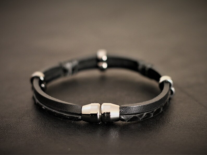 K&ouml;nnte beinhalten: Ein schwarzes Lederarmband mit einem Magnetverschluss und silbernen Akzenten.