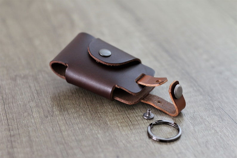 May include: A brown leather key holder with a snap closure and a metal key ring. The holder is open and the key ring is detached.