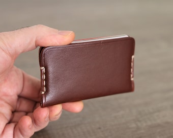 Può includere: Un portafoglio in pelle marrone con cuciture bianche. Il portafoglio è chiuso e viene tenuto in una mano.