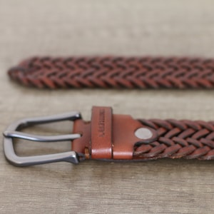 K&ouml;nnte beinhalten: Ein brauner geflochtener Lederg&uuml;rtel mit einer silbernen Schnalle. Der G&uuml;rtel hat das Wort "Leather" auf dem Riemen gepr&auml;gt.