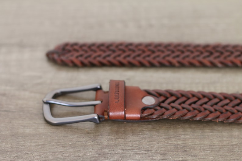 K&ouml;nnte beinhalten: Ein brauner geflochtener Lederg&uuml;rtel mit einer silbernen Schnalle. Der G&uuml;rtel hat das Wort "Leather" auf dem Riemen gepr&auml;gt.