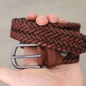 K&ouml;nnte beinhalten: Ein brauner geflochtener Lederg&uuml;rtel mit einer silbernen Schnalle. Der G&uuml;rtel hat den Text "Leather" gepr&auml;gt.