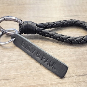 Op de afbeelding: Zwarte leren sleutelhanger met een gevlochten lus en een rechthoekig label met de tekst "EMILY & PAUL".