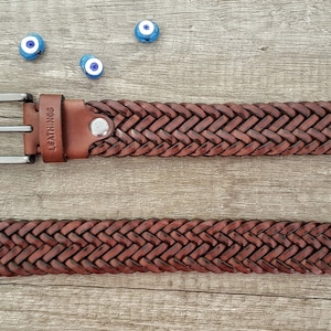 K&ouml;nnte beinhalten: Ein brauner Lederg&uuml;rtel mit geflochtenem Design und einer silbernen Schnalle. Der G&uuml;rtel liegt auf einer Holzoberfl&auml;che mit blauen und wei&szlig;en Gl&uuml;cksbringer-Augen um ihn herum. Der G&uuml;rtel hat das Wort "LEATHINGS" auf dem Leder gepr&auml;gt.