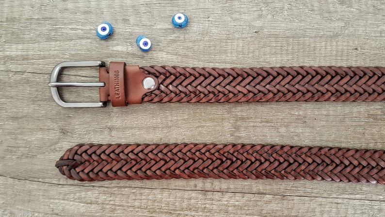 K&ouml;nnte beinhalten: Ein brauner Lederg&uuml;rtel mit geflochtenem Design und einer silbernen Schnalle. Der G&uuml;rtel liegt auf einer Holzoberfl&auml;che mit blauen und wei&szlig;en Gl&uuml;cksbringer-Augen um ihn herum. Der G&uuml;rtel hat das Wort "LEATHINGS" auf dem Leder gepr&auml;gt.