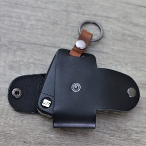 May include: Black leather key fob with a brown leather strap and a silver ring. The fob has a silver button and a small metal logo.
