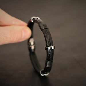 K&ouml;nnte beinhalten: Ein schwarzes Lederarmband mit silbernen Akzenten. Das Armband ist geflochten und hat einen einfachen Verschluss.