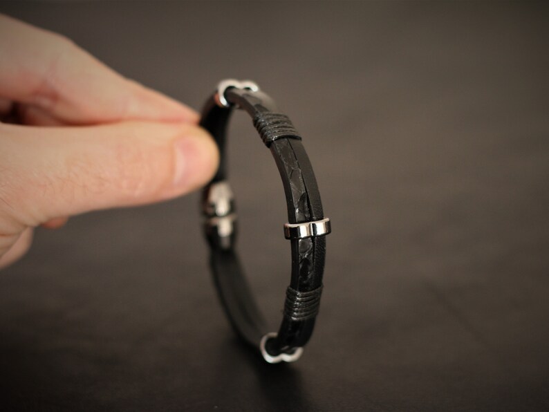 K&ouml;nnte beinhalten: Ein schwarzes Lederarmband mit silbernen Akzenten. Das Armband ist geflochten und hat einen einfachen Verschluss.