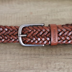 K&ouml;nnte beinhalten: Ein brauner geflochtener Lederg&uuml;rtel mit einer silbernen Schnalle. Der G&uuml;rtel hat das Wort "LEATHER" auf dem Riemen gepr&auml;gt.