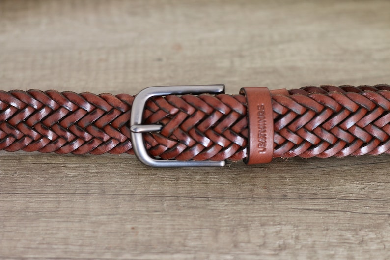K&ouml;nnte beinhalten: Ein brauner geflochtener Lederg&uuml;rtel mit einer silbernen Schnalle. Der G&uuml;rtel hat das Wort "LEATHER" auf dem Riemen gepr&auml;gt.