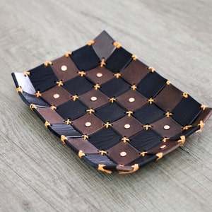 May include: A woven leather tray with a checkered pattern in black and brown. The tray has a decorative stitching detail and copper colored rivets.