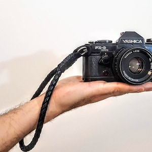 Könnte beinhalten: Eine schwarze YASHICA FX-D Quartz Kamera mit einem YASHICA ML 50mm Objektiv. Die Kamera wird in einer Hand mit einem schwarzen geflochtenen Band gehalten.