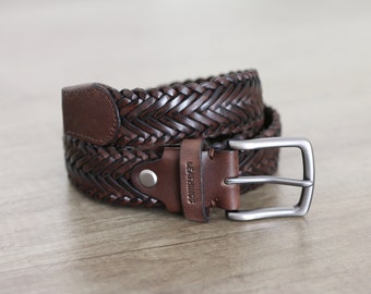 May include: A braided brown leather belt with a silver-colored buckle. The belt is rolled up, showcasing the intricate woven design. The leather has a rich, dark brown color, and the buckle is a simple, rectangular shape.