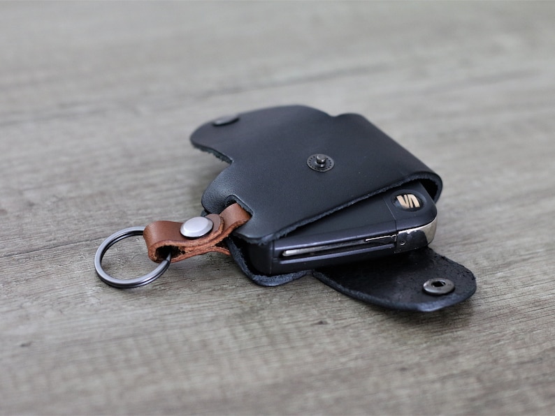 May include: A black leather key fob with a brown leather strap and a silver key ring. The fob has a flap that covers a key.