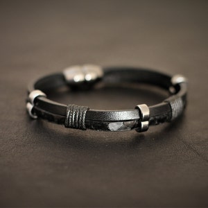K&ouml;nnte beinhalten: Ein schwarzes Lederarmband mit silbernen Akzenten. Das Armband hat ein geflochtenes Lederdesign und einen silbernen Verschluss.