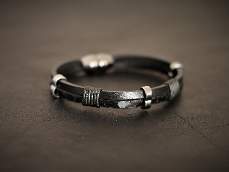 K&ouml;nnte beinhalten: Ein schwarzes Lederarmband mit silbernen Akzenten. Das Armband hat ein geflochtenes Lederdesign und einen silbernen Verschluss.