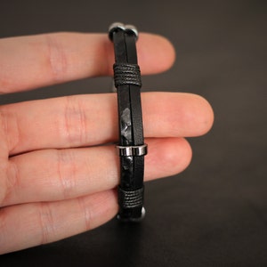 K&ouml;nnte beinhalten: Ein schwarzes Lederarmband mit silbernen Metallakzenten. Das Armband ist verstellbar und hat ein schlichtes Design.