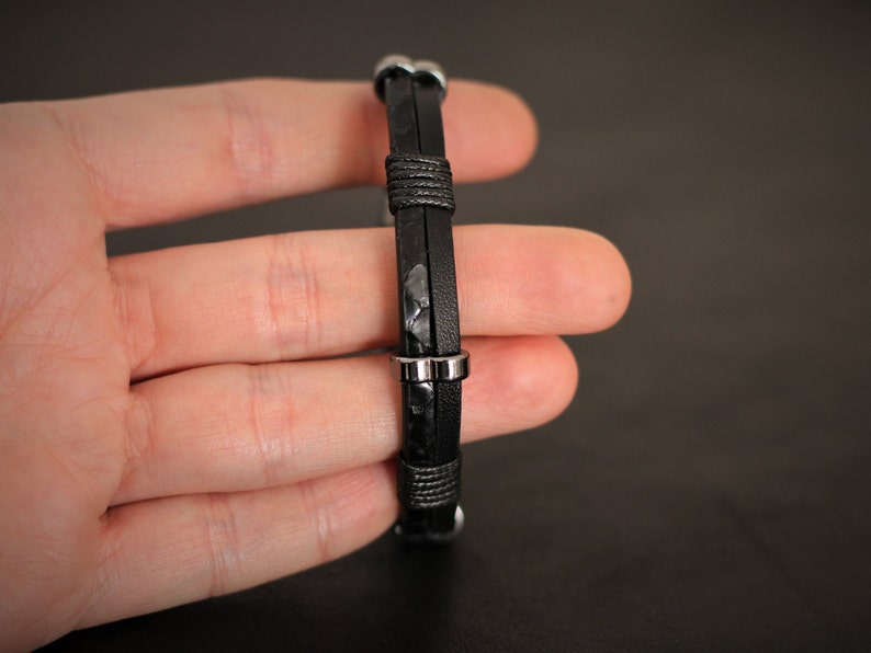 K&ouml;nnte beinhalten: Ein schwarzes Lederarmband mit silbernen Metallakzenten. Das Armband ist verstellbar und hat ein schlichtes Design.