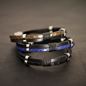 K&ouml;nnte beinhalten: Drei Lederarmb&auml;nder, die &uuml;bereinander gestapelt sind. Das obere Armband ist braun mit einem strukturierten Muster, das mittlere Armband ist schwarz mit einem blauen strukturierten Muster und das untere Armband ist schwarz mit einer glatten Oberfl&auml;che. Jedes Armband hat silberne Metallakzente.