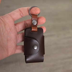 May include: A brown leather key holder with a metal ring and a brown leather strap. The key holder has a metal snap closure.