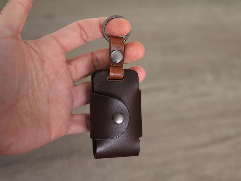 May include: A brown leather key holder with a metal ring and a brown leather strap. The key holder has a metal snap closure.