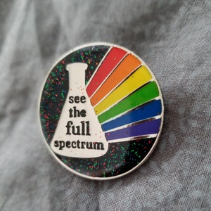 Puede incluir: Un pin de plata con un arcoíris y una probeta blanca con el texto "see the full spectrum".