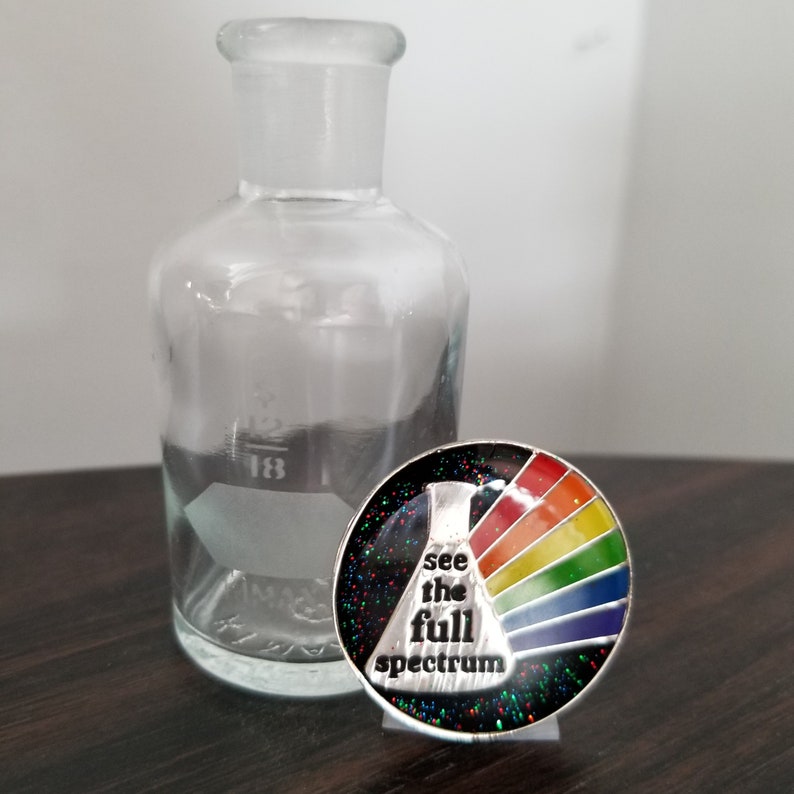 May include: A silver pin with a rainbow spectrum and a black background. The pin features a beaker with the text "see the full spectrum".