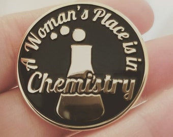 May include: A black and gold enamel pin with the text "A Woman's Place is in Chemistry" and a stylized beaker.