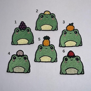 May include: Six embroidered frog patches are shown, each with a different fruit on its head. The fruits include grapes, a lemon, an orange, a mushroom, a pineapple, and a strawberry. The frogs are green with a light yellow belly and a black border.
