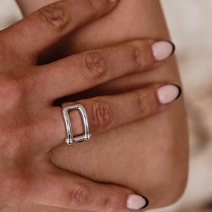 May include: A silver-colored ring with a rectangular design and two small ball accents. The ring is worn on a hand with manicured nails. The ring's design is a modern take on a classic style.