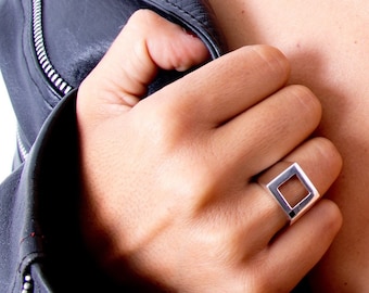 May include: A polished silver ring featuring a prominent square-shaped open frame design. The band is smooth and metallic, showcasing a clean geometric aesthetic that sits on the finger. The ring is worn alongside a dark leather jacket.