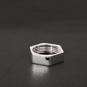 Könnte beinhalten: Ein silberfarbener, sechseckiger Ring mit Innengewinde und einem kleinen, dunklen quadratischen Detail. Der Ring ist vor einem dunklen Hintergrund platziert, der seinen metallischen Glanz und sein geometrisches Design hervorhebt.