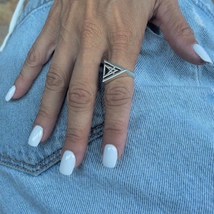 May include: A silver ring with a geometric triangle design. The ring is on a person's finger, and the person's hand is resting on a pair of blue denim jeans.