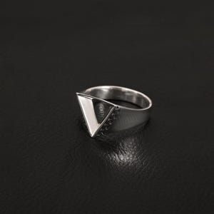 May include: A silver-toned ring featuring a prominent triangular face. The front surface of the triangle is smooth and reflective, while the sides of the triangular setting are accented with a series of small, dark, recessed circular details.