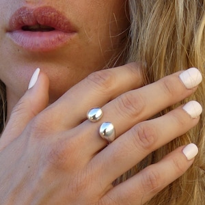 May include: A silver ring featuring two polished, rounded spheres. The ring is worn on a hand with light pink nail polish. The background shows a person's lips and blonde hair.