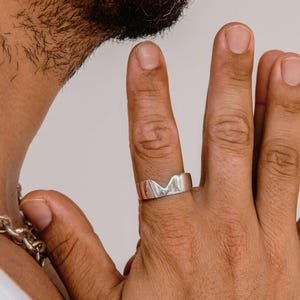 K&ouml;nnte beinhalten: Ein polierter Silberring mit einem markanten, bergf&ouml;rmigen Ausschnitt am Ringband. Der Ring wird am Mittelfinger getragen und zeichnet sich durch eine glatte, reflektierende Metalloberfl&auml;che und eine offene Silhouette aus.