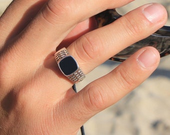 Könnte beinhalten: Ein silberfarbener Ring mit einer glatten, rechteckigen schwarzen Einlage in einem strukturierten Gliederband. Der Ring hat eine polierte Oberfläche und einen flachen, mittig platzierten schwarzen Stein, der einen Kontrast zum silbernen Metallband bildet.