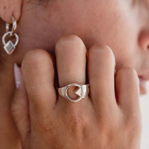Op de afbeelding: Een zilveren ring met een geometrisch ontwerp met een cirkel en een driehoek. De ring wordt aan een vinger gedragen en toont zijn gepolijste metalen afwerking. De sieraden maken deel uit van een collectie ringen en oorbellen.