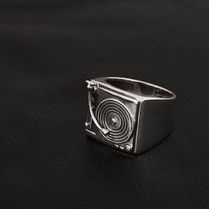 May include: A polished silver-toned ring featuring a detailed relief design of a turntable on its face. The ring displays a circular record platter with concentric grooves and a tonearm extending from the side, crafted in a solid metal band.