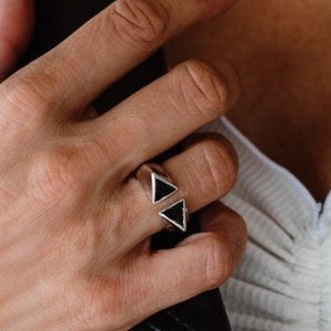 Puede incluir: Un anillo de plata con dos piedras negras en forma de triángulo. El anillo se lleva en el dedo medio de una mano.