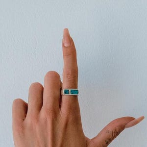 May include: A silver ring featuring a rectangular turquoise stone inlay set horizontally on a thin band. The ring is worn on the middle finger, showcasing a clean, metallic finish and a vibrant blue-green stone surface that sits flush within the setting.