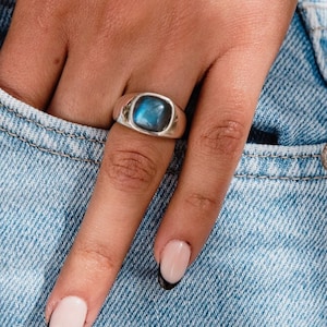 May include: A silver signet ring featuring a smooth, polished, square-shaped labradorite gemstone with a blue iridescent flash. The ring has a thick, solid band and is worn on the ring finger, showcasing the stone's natural color play.